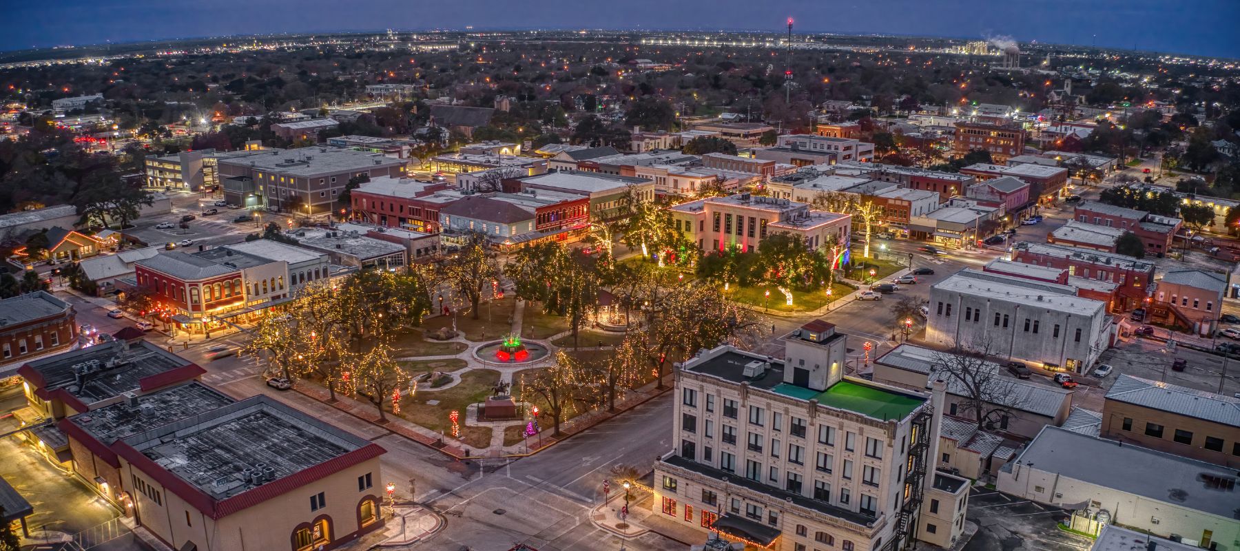 A festive aerial shot of downtown Seguin, Texas at night, with holiday lights illuminating the central park and historic buildings